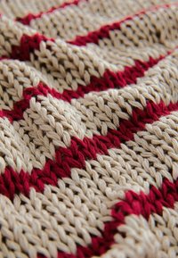 Gros plan sur un tissu tricoté avec des fils beige et rouge créant des motifs rayés diagonaux et des points texturés visibles.