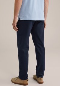 Marineblaue Hosen im Slim-Fit-Schnitt, aus glattem Stoff gefertigt. Kombiniert mit beigen Wildlederschuhen. Sichtbare Ziernähte und seitliche Taschen.