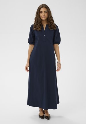 Femeie cu păr castaniu creț, care poartă o rochie lungă albastru închis cu mâneci bufante până la cot și pantofi negri cu vârf ascuțit, stând în fața unui fundal simplu.