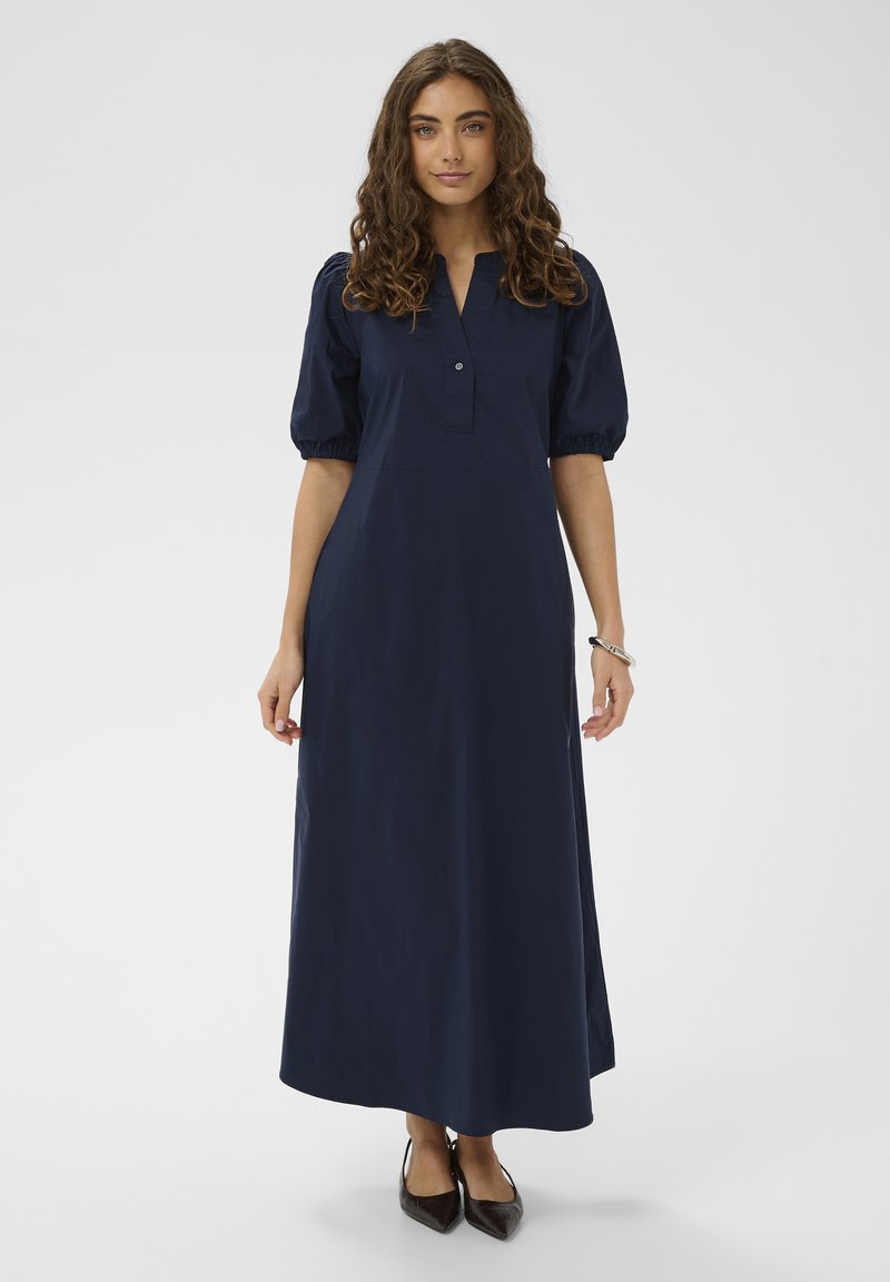 Žena s kovrčavom smeđom kosom, odjevena u tamnoplavu dugu haljinu s naboranim polurukavima i crnim šiljastim cipelama, stoji nasuprot jednobojnoj pozadini.