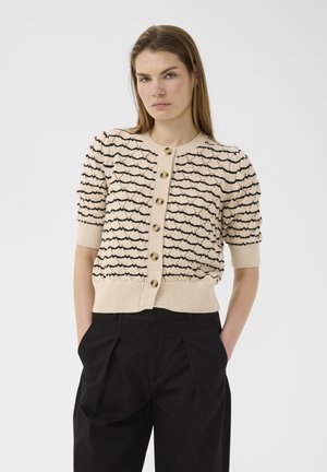 Frau trägt eine beige kurzärmelige Strickjacke mit schwarzem Wellenmuster und Knöpfen, kombiniert mit hoch taillierten schwarzen Faltenhosen.