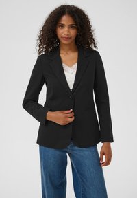 Göndör hajú nő, fekete blézerben, fehér csipkés top és kék farmernadrág felett, egy sima világos háttér előtt áll.