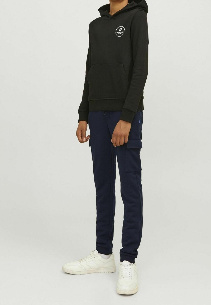 Personne portant un sweat à capuche noir avec un logo, un pantalon cargo bleu marine et des baskets blanches, se tenant devant un fond clair.