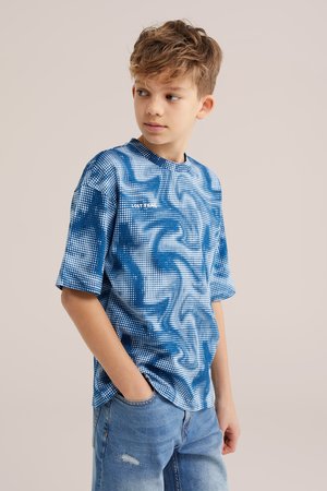 Fant z razmrščenimi lasmi, ki nosi kratko majico z modro-belim vzorcem in svetlo modre kavbojke, gleda v stran z eno roko v žepu.