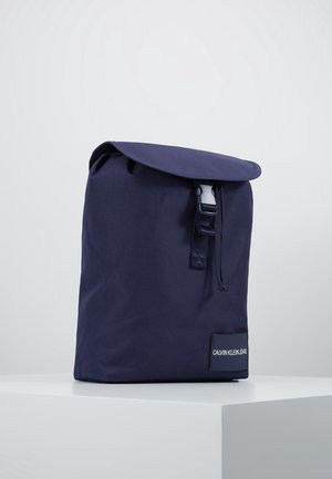Rygsæk i navyblåt stof med klap og spænde, med en "Calvin Klein Jeans"-etiket på det nederste forreste hjørne.