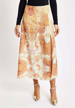 Long skirt featuring an orange and cream paisley pattern, made of lightweight fabric with a side slit and paired with gold heeled shoes.