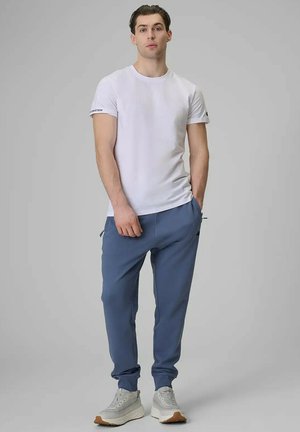 Młody mężczyzna stoi z rękami w kieszeniach, ubrany w białą koszulkę z krótkim rękawem, niebieskie spodnie dresowe i szare trampki na szarym tle.