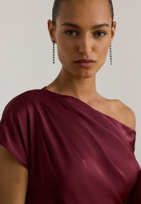 Vestido de cetim bordô com um ombro só, design drapeado; modelo usa brincos longos pendentes de esmeralda, textura lisa e brilho suave.