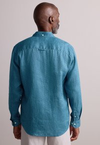 Camisa de manga larga turquesa con un patrón geométrico, puños con botones y detalle de lazo en la parte posterior para colgar. Textura de tela suave.