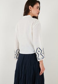Blusa de lino blanca con mangas hasta el codo que presenta bordados geométricos azules en los puños. Combinada con una falda plisada azul marino.