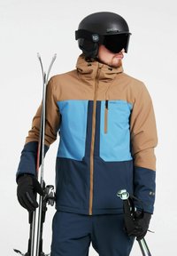 Chaqueta de esquí en marrón, azul y marino con un bolsillo en el pecho con cremallera. Se lleva con guantes negros, sosteniendo esquís. Acabado mate y diseño aerodinámico.