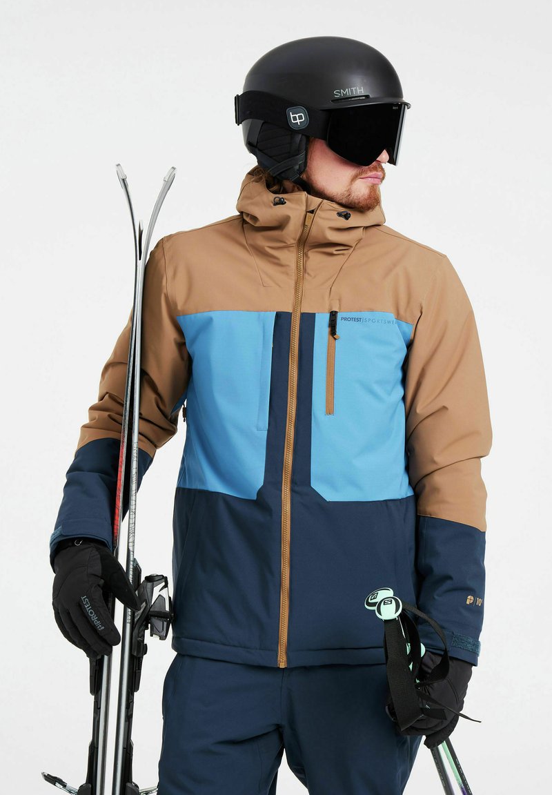 Chaqueta de esquí en marrón, azul y marino con un bolsillo en el pecho con cremallera. Se lleva con guantes negros, sosteniendo esquís. Acabado mate y diseño aerodinámico.