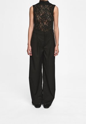Femme portant un haut en dentelle noire sans manches et un pantalon noir taille haute à jambes larges, debout sur un fond blanc.