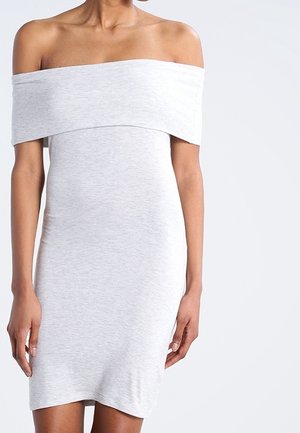 Frau in einem hellgrauen, schulterfreien, taillierten Kleid, steht mit entspannten Armen an den Seiten vor einem einfarbigen hellen Hintergrund.