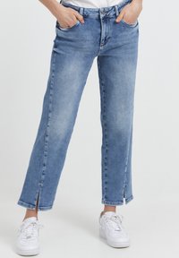 Mujer con jeans cortos de color azul claro con aberturas en los tobillos y zapatillas blancas, con las manos en los bolsillos, de pie contra un fondo blanco.