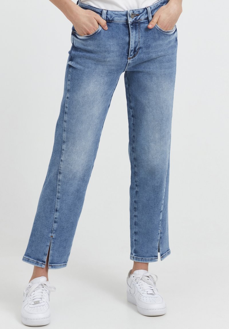 Mujer con jeans cortos de color azul claro con aberturas en los tobillos y zapatillas blancas, con las manos en los bolsillos, de pie contra un fondo blanco.