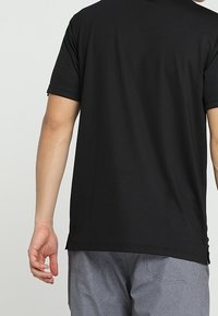 Mies pukeutuneena yksiväriseen mustaan lyhythihaiseen paitaan ja harmaisiin housuihin, kuvattuna selin, vasen käsi rentona sivulla.