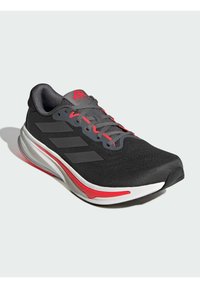 Black athletic shoe with red accents, mesh upper, grey laces, three black stripes on side, and a textured sole for grip.