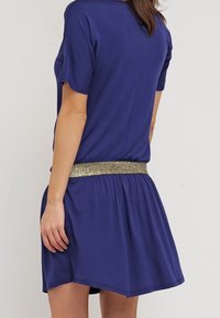 Lyhythihainen violetti mekko, jossa on teksturoitu kultainen kimaltava vyö, rento istuvuus ja kerätty vyötärö, päättyen polven ylle.