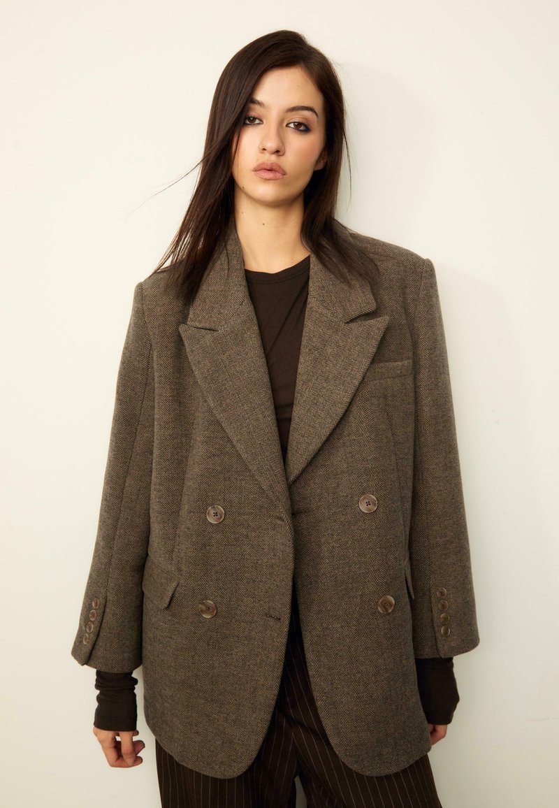 Rjav herringbone dvoredni blazer z velikimi gumbi in strukturiranimi rameni, nosi se čez temno bluzo, ki ima dolge rokave.