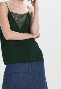 Grön camisole med spetsdetalj vid V-ringning, justerbara spaghettiband och en lös passform, tillsammans med en jeanskjol. Slät textur.