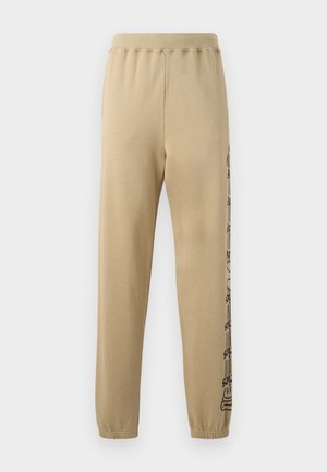 Beigās sporta bikses ar brīvu piegriezumu, elastīgu jostu un sašaurinātām piedurknēm. Uz kreisās kājas ir melnas grafikas. Izgatavotas no mīksta auduma.