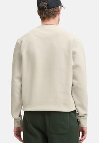Sweat-shirt beige clair avec col rond, poignets côtelés, et détail de selle dans le dos, porté avec un pantalon vert foncé. Texture douce et coupe décontractée.