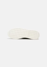 Semelle d'une chaussure en caoutchouc blanche avec un motif à grille, vue à plat sur un fond uni gris clair.