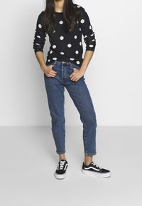 Laurakansallinen neule, jossa on valkoisia pilkkuja, yhdistettynä vaaleansinisiin korkeavyötäröisiin farkkuihin ja mustiin lenkkareihin, joissa on valkoisia yksityiskohtia.