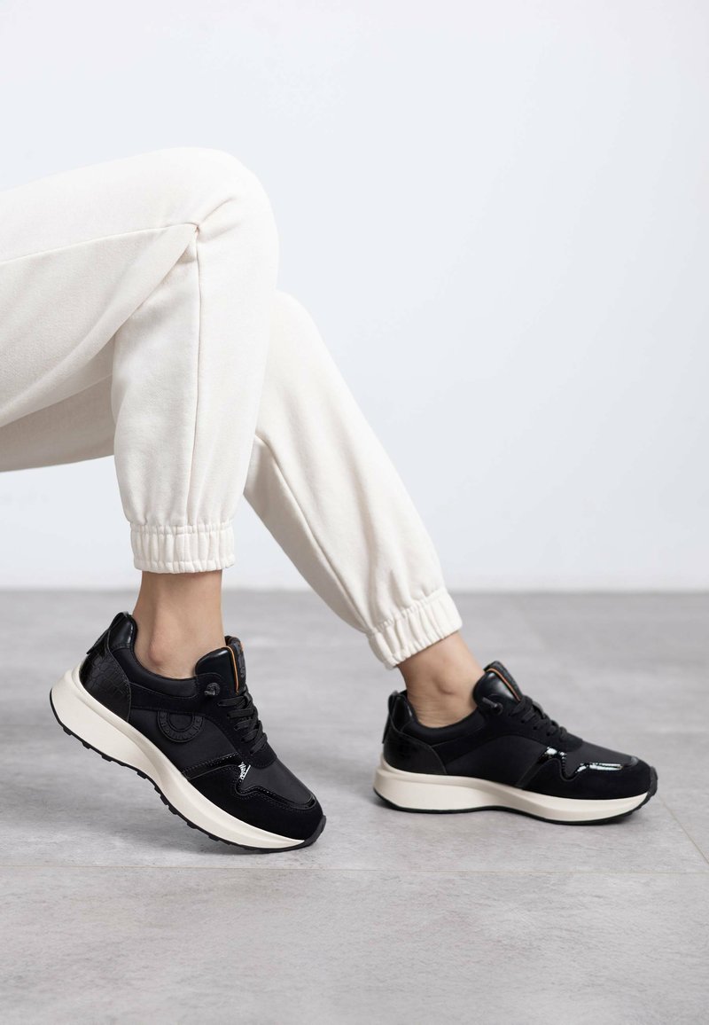 Zapatillas deportivas negras con una textura suave, que cuentan con una suela gruesa de color crema y acentos negros en contraste. Diseño con cordones y detalle del logo.