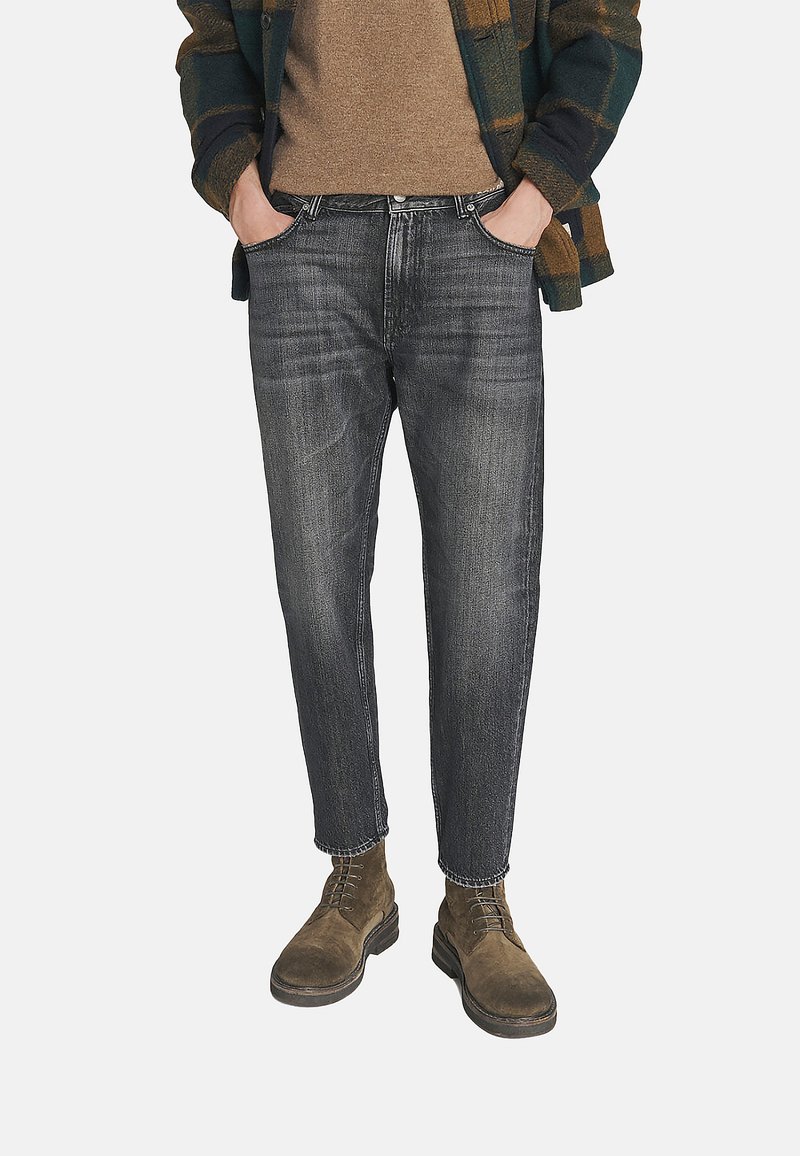 Uomo con jeans neri scoloriti, stivali in suede marroni, maglione marrone e giacca a quadri verde scuro e marrone, con le mani in tasca.