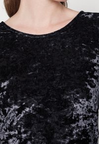 Haut en velours noir avec une surface douce et texturée, présentant un large décolleté et une finition légèrement brillante.