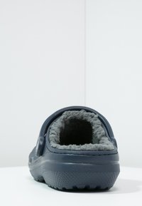 Chausson bleu marine avec une douce doublure en fourrure grise et une semelle en caoutchouc texturée. Il présente un dos rond et un design à enfiler bas.