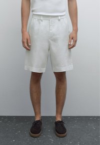 Shorts en lin blanc avec une devant plat, une taille élastique et des poches latérales. Le tissu a une apparence légère et texturée.