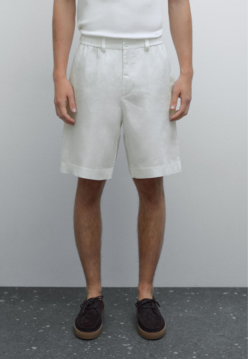Shorts en lin blanc avec une devant plat, une taille élastique et des poches latérales. Le tissu a une apparence légère et texturée.