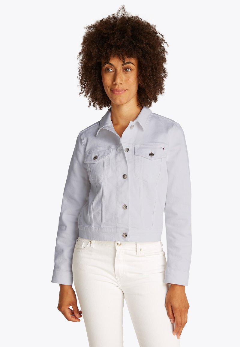 Woman with curly hair wearing a light gray buttoned jacket and white pants standing against a plain background.