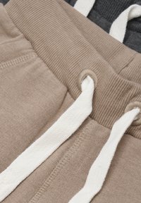 Pantalons de jogging en coton beige et gris foncé. Présente une taille côtelée avec des cordons de serrage blancs et des détails de couture visibles.
