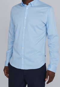 Jasnoniebieska koszula z długim rękawem, o gładkiej fakturze i zaokrąglonym dole. Białe guziki i dopasowany krój. W połączeniu z ciemnymi spodniami.