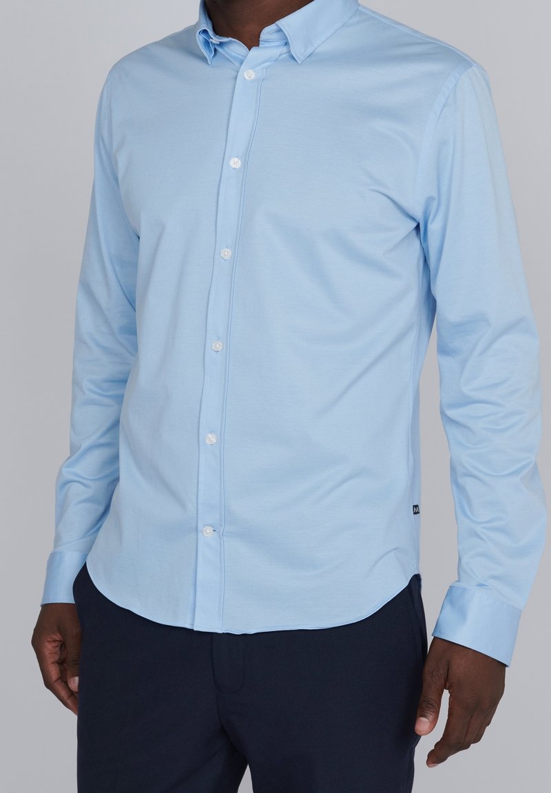 Jasnoniebieska koszula z długim rękawem, o gładkiej fakturze i zaokrąglonym dole. Białe guziki i dopasowany krój. W połączeniu z ciemnymi spodniami.