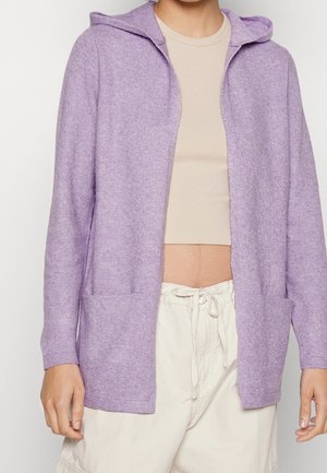 Personne portant un cardigan à capuche lilas sur un top court beige, associé à un pantalon taille haute beige clair avec cordon de serrage.