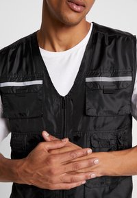 Hombre con chaleco utilitario negro con franjas reflectantes sobre camisa blanca, con las manos entrelazadas a la altura de la cintura sobre un fondo liso.