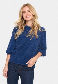 Blå, fluffig tröja gjord av mjukt material med en avslappnad passform, med trekvartsärmar och rund halsringning. Bärs med mörka jeans.