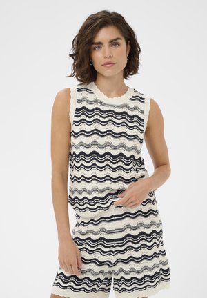 Femme aux cheveux courts bruns portant un haut sans manches crème et un short assorti avec des motifs de rayures ondulées noires sur un fond blanc.