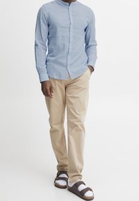 Camicia azzurro chiaro a maniche lunghe con colletto a bande, chinos beige e sandali marroni con calzini bianchi, su uno sfondo bianco semplice.