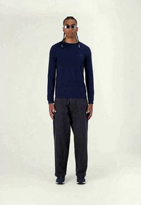 Tengerészeti kék hosszú ujjú póló, mellkason logóval, sötét cargo nadrággal párosítva. A modellen napszemüveg és sportcipő található. Simább textúrájú, laza szabású.