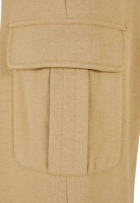 Beżowy materiał odzieżowy z dużą, płaską kieszenią z klapą, przeszytymi brzegami i miękką teksturą; zaprojektowany z myślą o funkcjonalności i komforcie.