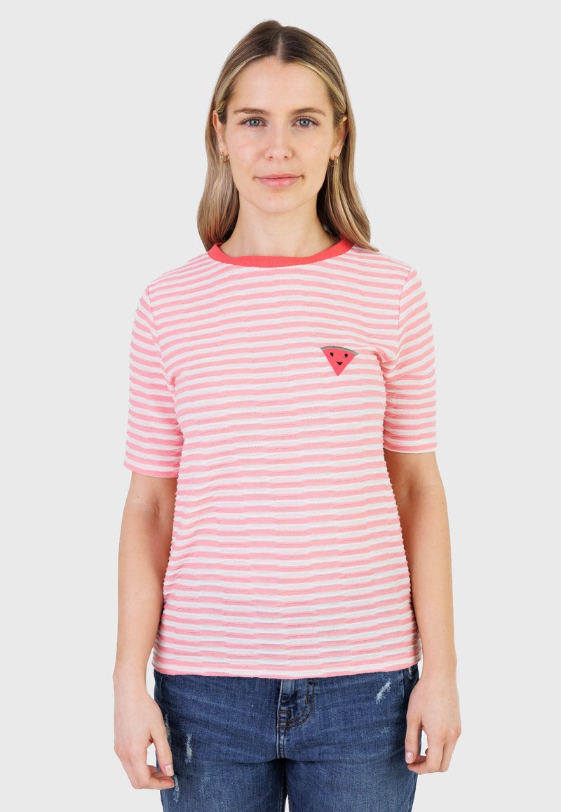 Femme aux cheveux blonds raides portant un t-shirt rayé rose et blanc avec un petit écusson de pastèque rouge et un jean bleu.