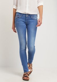 Persona que lleva jeans ajustados de color azul claro, una camisa blanca con mangas arremangadas y sandalias metálicas de doble hebilla sobre un fondo sencillo.