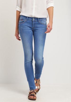Persona que lleva jeans ajustados de color azul claro, una camisa blanca con mangas arremangadas y sandalias metálicas de doble hebilla sobre un fondo sencillo.