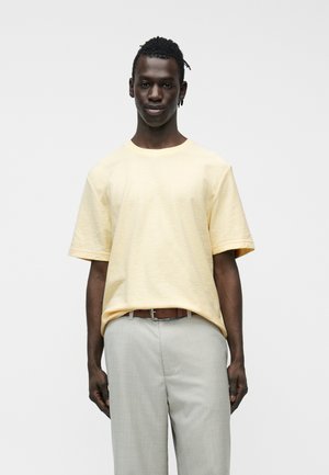 Mand med kort, krøllet hår iført lys gul kortærmet skjorte stoppet ned i lysegrå bukser med brunt bælte, stående på hvid baggrund.